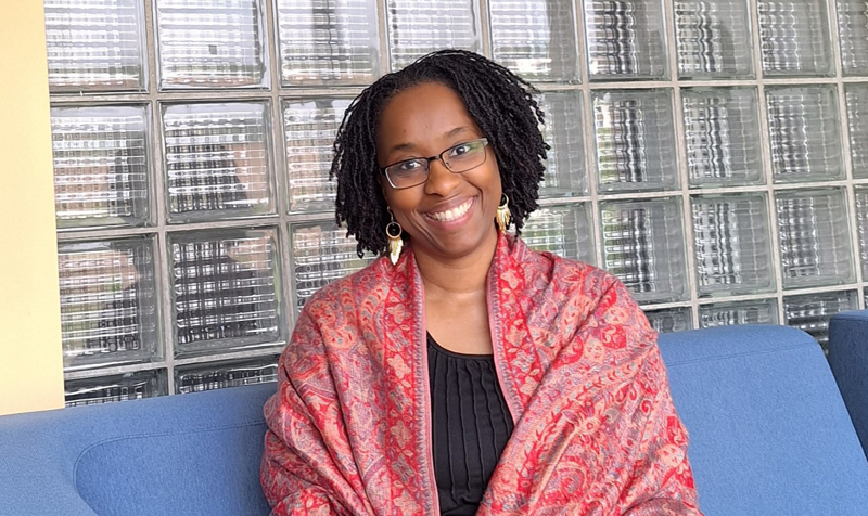 A young professional woman wearing a red shawl sits, smiling on a blue sofa, with glass block wall behind.