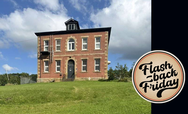 A 2 story red brick building set against a field of green grass and a blue sky.
