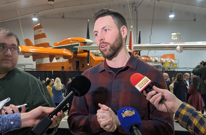 A man is taking questions from reporters in an aircraft hangar with a water bomber aircraft in the backgroung