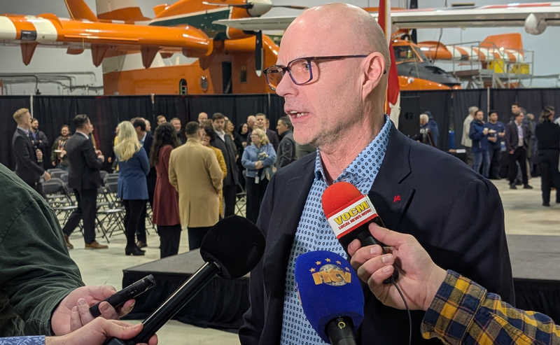 A man, (Neil Chaulk) takes questions from reporters, in an aircraft hangar, with an airplane in the background