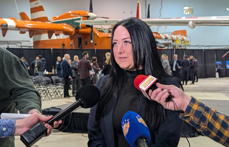 A woman, Falon O'Keefe, takes questions from reporters in an aircraft hangar with an airplane in the. background