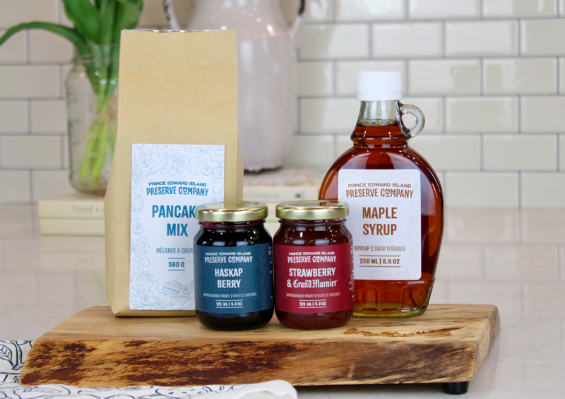 4 PEI Preserve Company products displayed on a wooden charcuterie type board. from left to right are: a brown paper bag of Pancake mix, 2 small jars of jam... one called Haskap Berry and the other is Strawberry and Grand Marnier, and then a glass bottle of maple syrup.