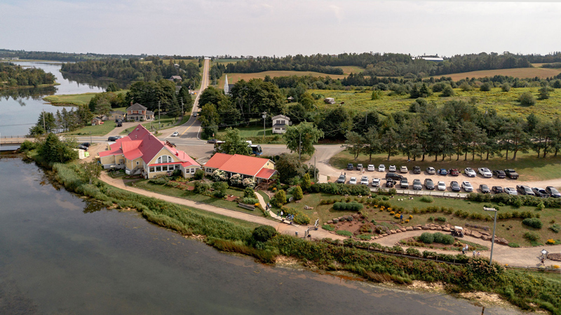 A view from above of the PEI Preserve Company. The pale yellow, 2 storey, wood siding building is at the edge of a body of water, and amidst grassy areas and a walking path. Also showing the parking lot full of cars, presumably customers.