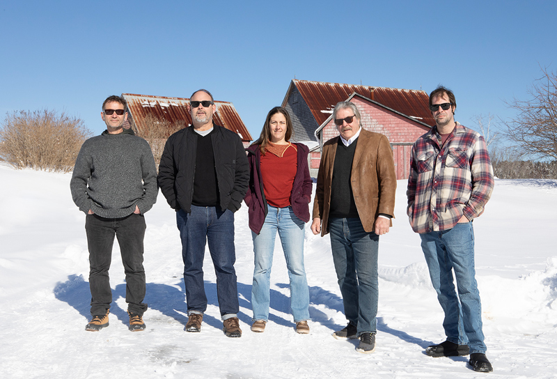 a team of 5 people (4 men, 1 woman-in the middle) standing in the snow with barns in the background