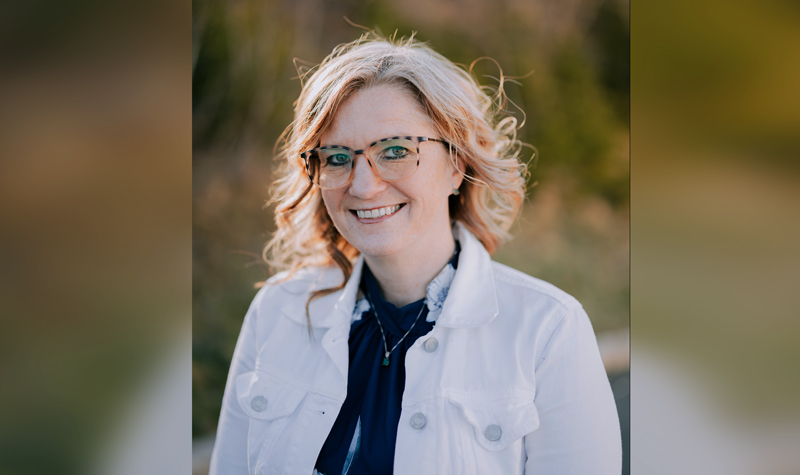A young woman with shoulder length blond hair, wearing a white casual jacket over a navy blouse, photographed against a background of greenery