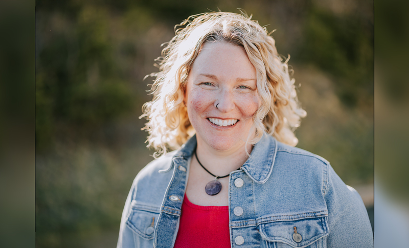 A smiling young woman with curly blonde hair, wearing a light denim jacket over a red blouse, photographed against greenery