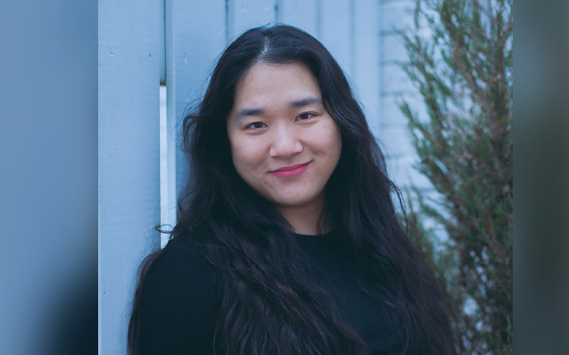 A young smiling Asian woman with long hair and a dark jacket, photographed against a white picket fence