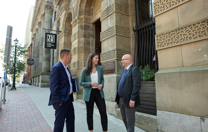 from left to right: a man, a woman, and another man, business casual, chatting outdoors in front of a historic looking stone commercial building