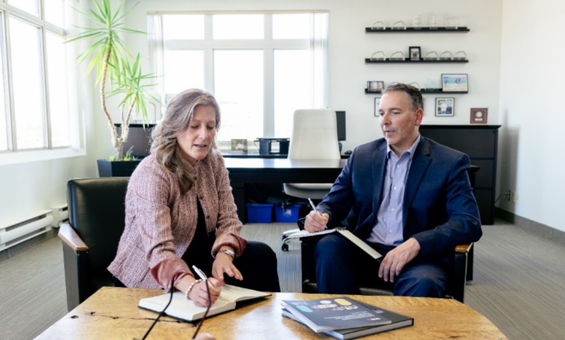 a woman and a man, dressed in business casual, in a meeting with 2 people at the near side of the table partly shown, indicating a casual business meeting, in a large, bright and modern office