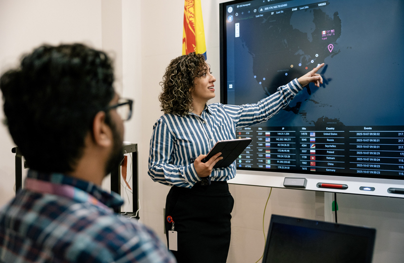 a young professional businesswoman in front of a smart board screen, pointing and explaining to a seated young professional man watching and listening to her