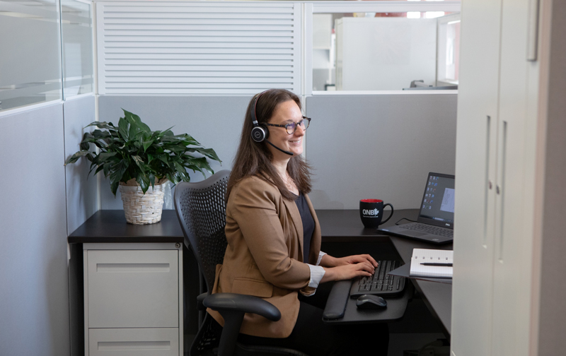 a smiling business professional woman seated at a desk and computer, wearing a headset, in a modern business cubicle