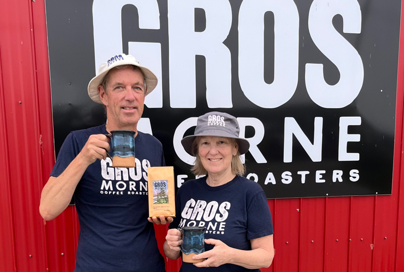 a man and a woman standing in front of a red building's sign that says Gros Morne Coffee Roasters. They are wearing tshirts and hats with the same words on them, and are each holding a cup of coffee.