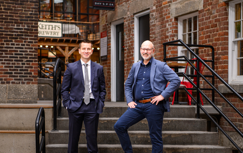 2 businessmen smiling, posed standing on stairs outside a red brick building.