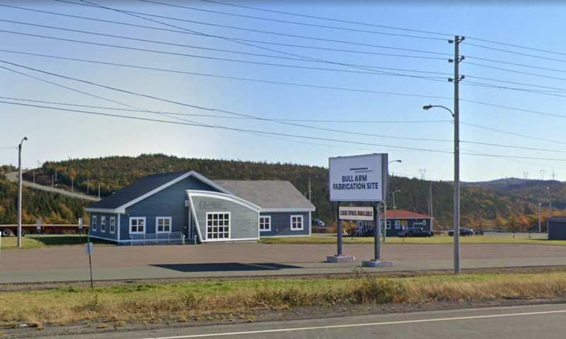 a one story building with blue siding and a sign that says Bull Arm Fabrication Site, against a background of a road and forest, and a highway in the foreground