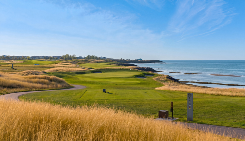 A golf course on the sea coast, with green grass, and wild grasses, set against a sunny day and smooth seas
