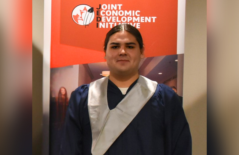 A young man is standing in front of an orange banner, smiling, and wearing a graduation gown