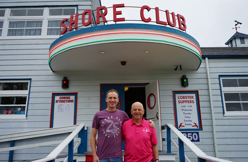  A middle aged man in jeans and a casual t shirt standing close to his father, dressed in jeans and a golf shirt, on the steps of a wooden building housing the Shore Club