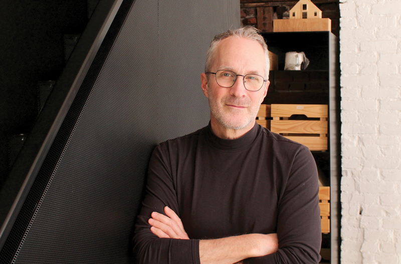 a middle aged, smiling man leaning against a wall of an interior renovation area, arms folded, wearing wire rimmed glasses, close cut greying beard, and wearing a brown turtle neck lightweight sweater