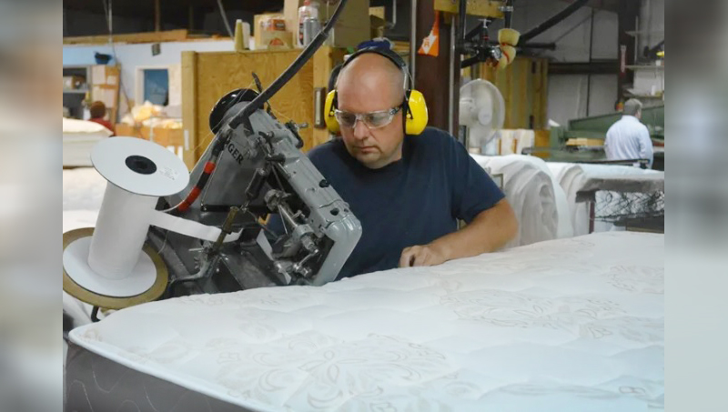 A man seated at an industrial sewing machine, wearing ear protection, and sewing a new mattress.