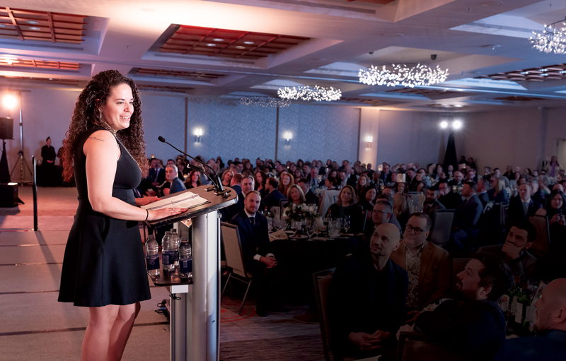  A smiling woman wearing a black dress, and with long dark curly hair, is standing at a podium in front of a large crowd of people seated at tables at a business event or conference.