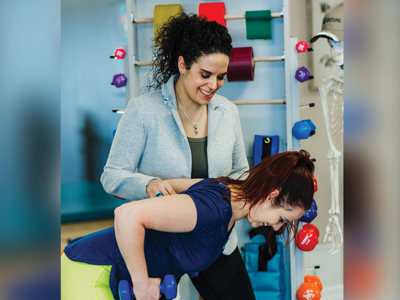A physiotherapist—a smiling young woman with curly dark long hair pulled back in a ponytail, is working with a client or patient, who is lifting weights while leaning forward, supported by the physiotherapist. In the background are various weights and resistance bands and other equipment, all organized very neatly and orderly.