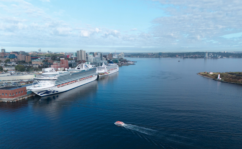 a very large cruise ship is docked in downtown Halifax, on a calm sunny day. There is a smaller cruise ship docked behind the large one.