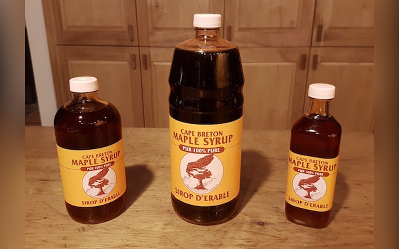 three bottles of Cape Breton Maple Syrup are on a wooden counter top with wooden door cupboards in the background. From left to right, the bottles are medium size, large, and small. The label has the map of Cape Breton, and a maple tree.