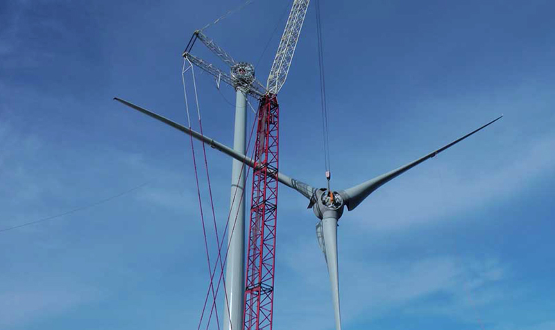 a large wind turbine is being assembled, with a large crane hoisting the piece that contains the 3 blades. This is a view from the ground, and is set against a blue sky. No visible indication of time of year or location.