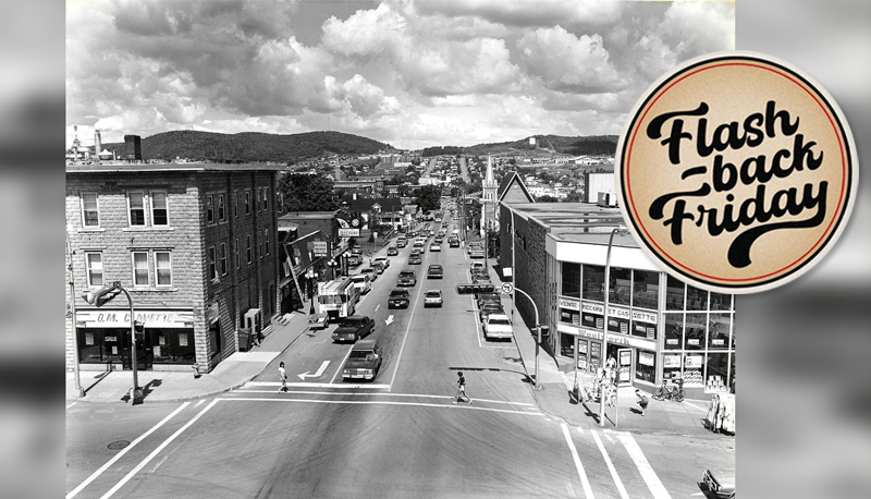 a view from above of a small city's downtown in the late 70s or possibly early 80s, showing an intersection with cars of the period at traffic lights, and pedestrians crossing , wearing summer clothing. The names of the the 2 businesses on the street corners are O.M. Clavette at the left and Woolworth at the right. The road between them recedes into the distance and has businesses and a church 