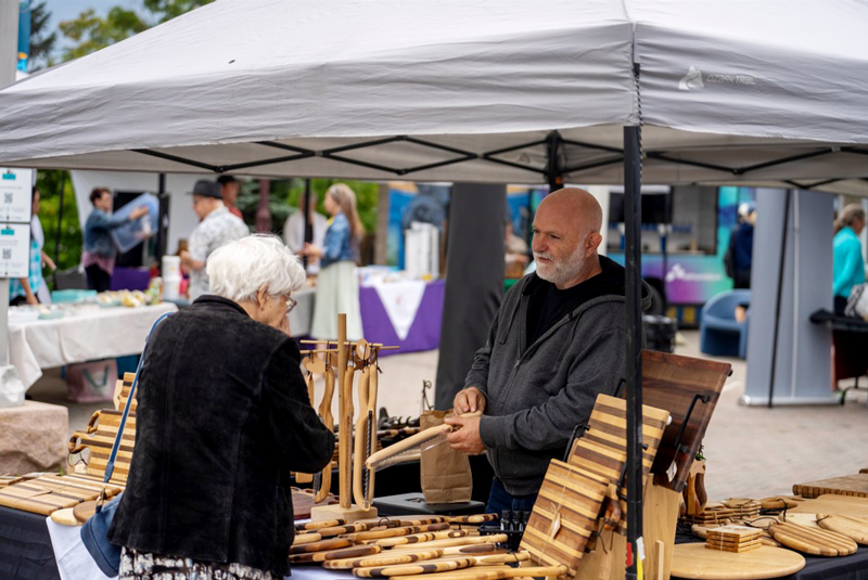 a vendor at an outdoor farmer's market talks to a customer about his hand crafted wood products, of which you can see cutting boards and bread knives. The woman appears older and has bright white hair, and the vendor is wearing a grey zip up hoodie over a black t shirt, and is bald with a grey mustache and beard. Other market customers can be seen behind this vendors stall which has a light grey canvas tent kind of roof.