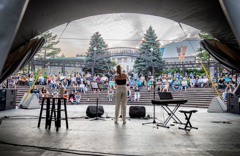viewed from behind backstage and looking through the arc shaped stage opening at an outdoor concert a woman is at a microphone, either speaking or singing. There are stands or seating in the form you would find in an amphitheatre and about half the capacity is taken up with concert goers apparently dressed for a possibly cooler summer day. 