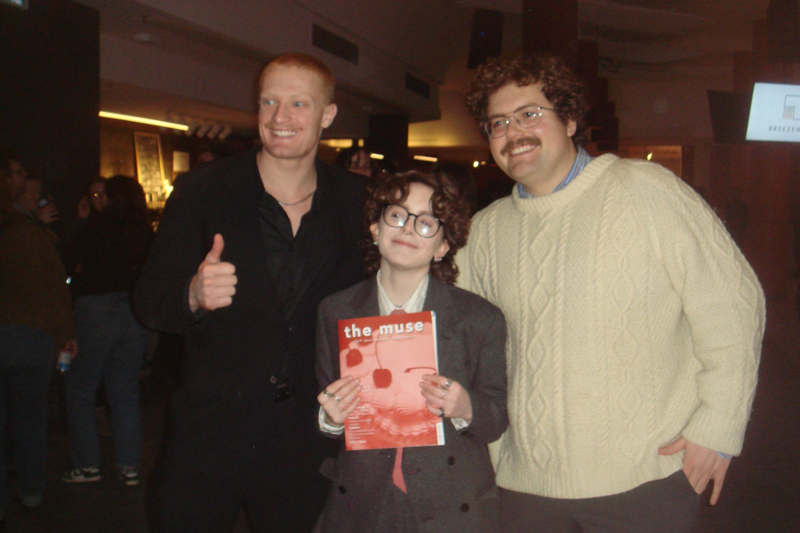 a group of 3 smiling university students: 2 taller and then a shorter person standing in front of them holding a copy of the publication, The Muse. The person left/back has short red hair and is dressed in a black button up shirt and giving the thumbs up sign; the other in the back has curly short-ish hair and a moustache, and is wearing glasses, a cream coloured knit sweater over a denim collar shirt; the person in fron also has shorter brown curly hair is wearing dark rimmed glasses and a white button up shirt under a brown or grey blazer, and also wearing several rings, and a pair of earrings. (the photo is a little blurry so it is is hard to see the details)