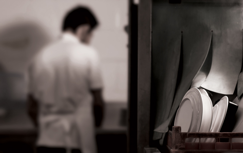 a man with dark hair and wearing a white short-sleeved shirt, and a long white apron, is back on to us and is washing dishes in what appears to be a restaurant kitchen. This scene is a little blurred, and at the right are dishes on a rack.