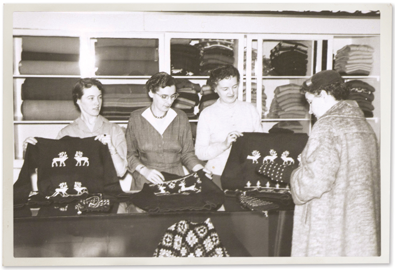 an old black and white photo from either th 1950s or early 60s, showing 4 women in the NONIA shop, looking at knit sweaters with patterns showing either moose or caribou. There are display cases behind them full of knitted goods.