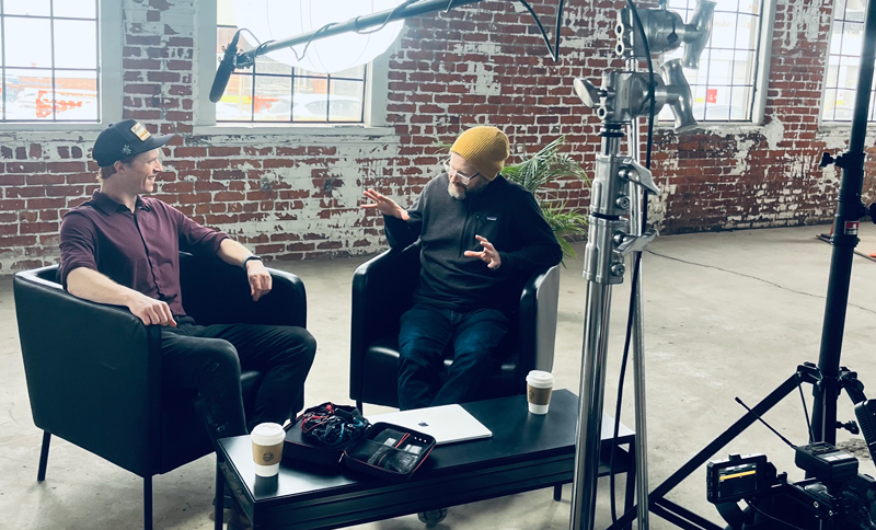 2 men seated in black club style chairs with recording equipment over them and in front. They are dressed casually... one in a button up shirt with sleeves rolled up, and the other in a fleece half-zip and a toque. The second man is wearing glasses, and gesturing during a conversation. The walls behind them are red brick with patchy white paint, and the windows appear to be that of an old factory setting.