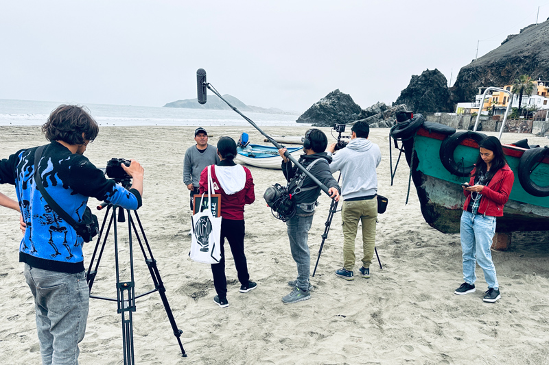 a film crew stands on a white sandy beach on a grey day with old fishing boats on the sand, and the ocean in the distance, with a rocky outcrop at the end of the beach. One of the crew is using a camera on a tripod, another is holding the mic boom and wearing a gear pack. Another appears to be overseeing the operation, and one is off to the side using a smartphone. They are all dressed casually for a cool day, wearing either sweaters or jackets left open.