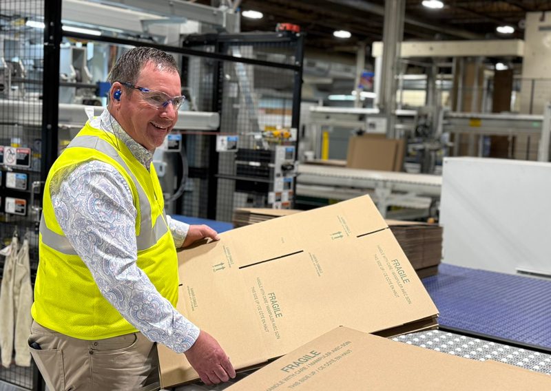 a man wearing khakis and a light grey paisley pattern button up shirt, stands smiling and looking off to the right. He is in a factory and handling just manufactured cardboard boxes. He is also wearing ear and eye protection.