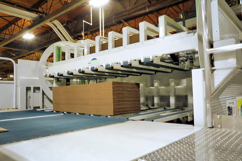 In a factory setting, a large machine is moving a big stack of newly manufactured brown cardboard boxes.