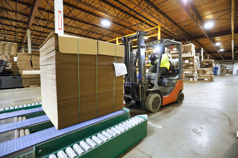Inside a really clean and tidy factory warehouse, a forklift driven by a worker in a yellow vest is picking up a pallet load of newly manufactured cardboard boxes.