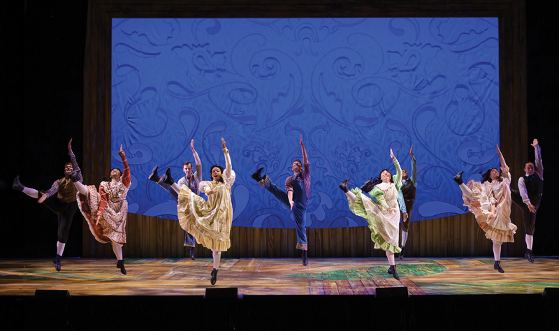 a group of 8 dancers, 4 women at the front of a theatre stage and 4 men further back. They are all dressed in costume appropriate to the Musical Anne of Green Gables. Each of them has their right leg kicked high and to their right, and their left arm raised above their heads. They appear to also be mid-jump. 