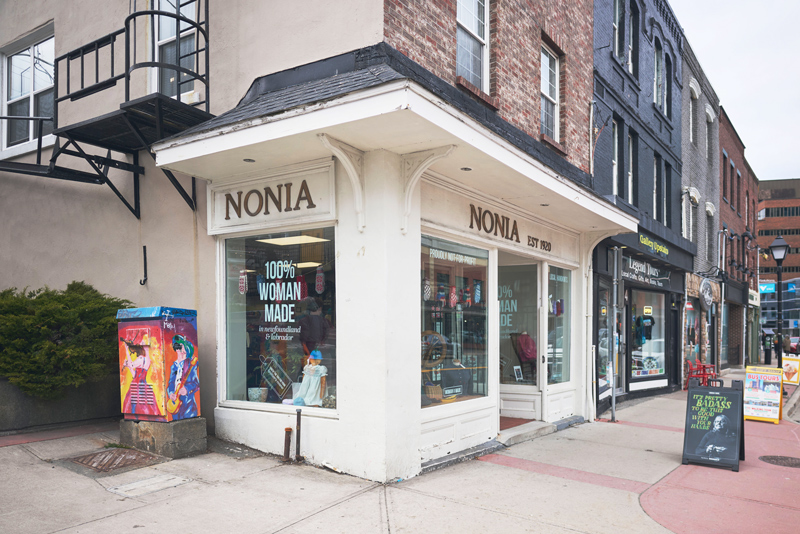 a store front on an old downtown city street, with large windows and the name NONIA on the front and on the side (the store is at the end of a row of connected buildings), and through the windows can be seen many colourful knit goods. On one window it says '100% Woman Made'. Above the level of the store, which is painted cream colour and has an overhang shingled roof supported by wooden curved support beams, the building front is red brick