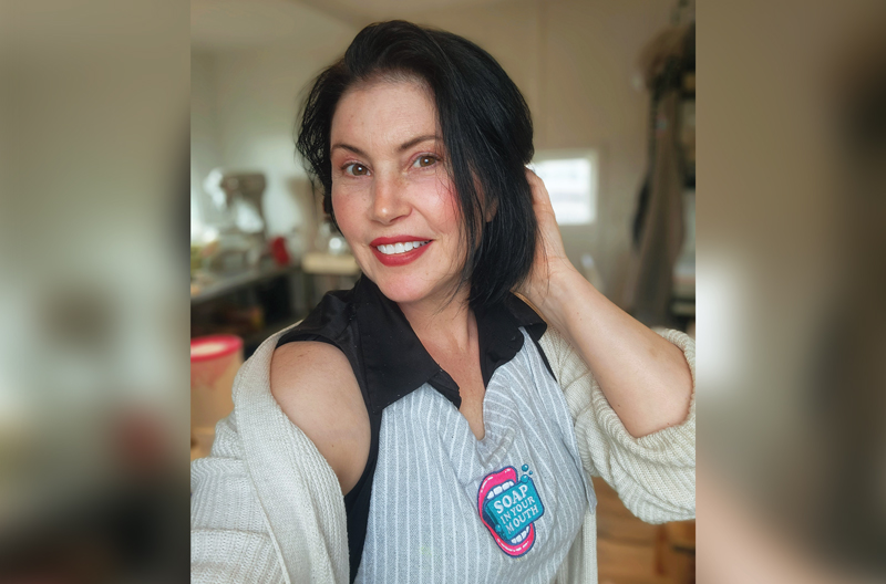 a young looking middle aged woman with long-ish dark hair and dark eyes, wearing lipstick, blush and with grooms brows, has her left hand on her hair behind her head. She is smiling, and is wearing a loose fitting cardigan over a sleeveless polo shirt with an apron over it. The apron has her business logo on it, which says "Soap in Your Mouth".