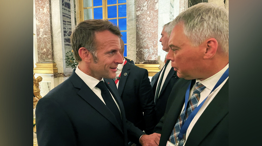 A relatively close up image of 2 men shaking hands, with a coupel of other people in the background amidst the opulant setting inside the Palace of Versailles. The man on the left has dark but greying hair and long sideburns, and the man on the right has very short almost white hair. They are both dressed in formal business suits