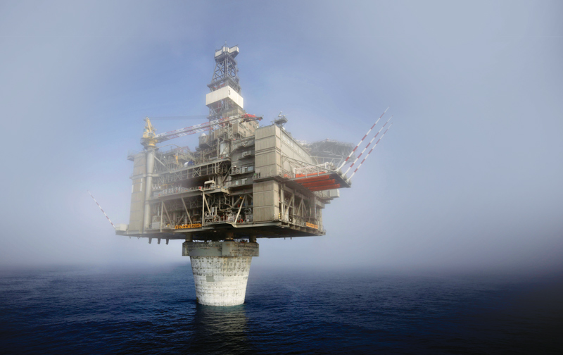 An oil drilling platform in the north atlantic ocean. The platform is high above the ocean surface, on top of a masive cylindrical concrete structure The sky is blue and the ocean is calm.
