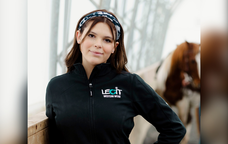 A young lady with dark eyes and long brown hair held back by a headband smiles at the camera, while a horse stands in the background. They are inside a stables or similar building. She is wearing a black zip-up fleece with the words LEGIT Western Wear on the upper left near the shoulder.