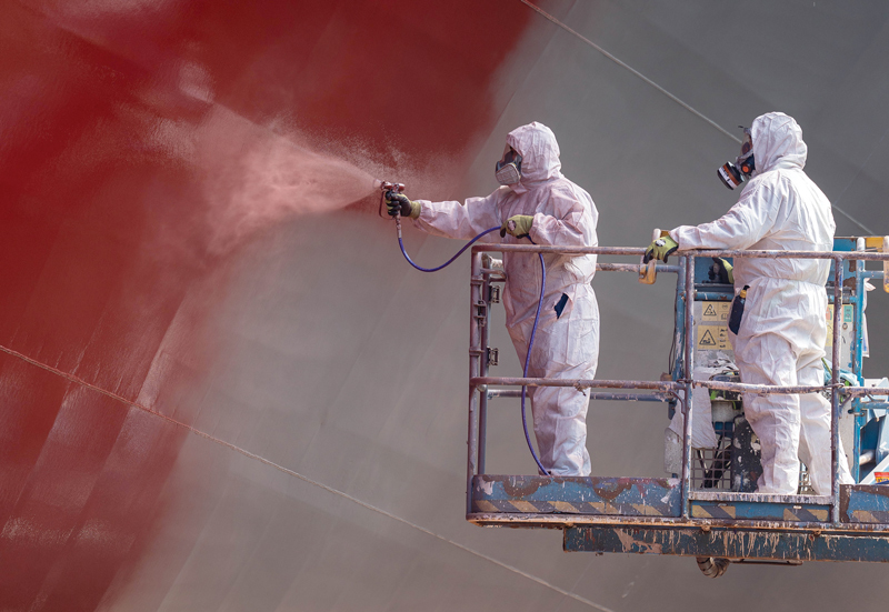 Two people dressed in protective gear are on an elevated platform and are using a spray gun to apply a red substance to the bare metal surface of a large ocean going vessel.