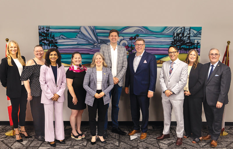 a group of smiling people, all dressed in business / business casual attire.The are standing against a wall with a large piece of artwork of a landscape in a sort of stained glass style.