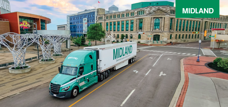 a Green Tractor trailer is pulling a white trailer with the word 'MIDLAND' on the side through a city street, with a historical appearing building in the background on the corner of the street. at the left are 3 modern sculptures that look like stylized trees. The word 'Midland' is on a small green rectangle as a separate graphic.