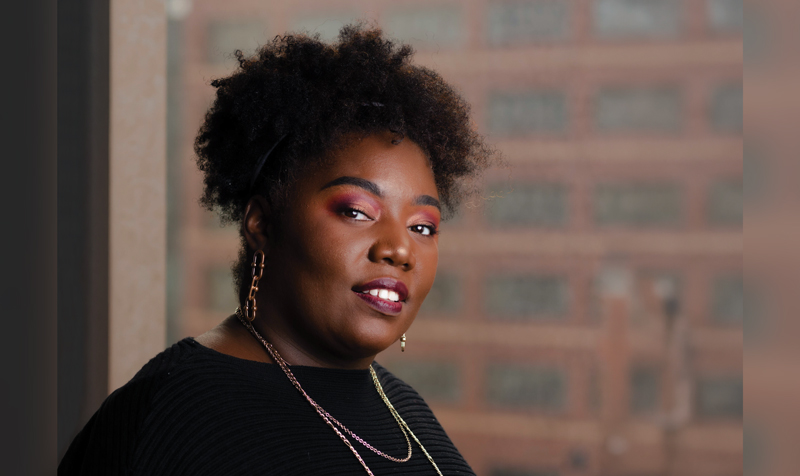 A portrait of a businesswoman in her braiding lounge salon. She is wearing a black sweater and long understated gold chain necklaces. She is against a background of an interior brick wall.