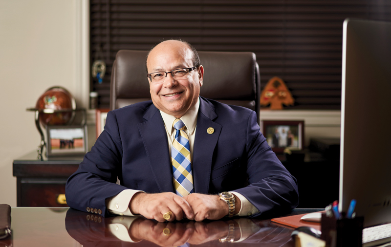 a businessman seated at his desk is smiling and looks happy. He is wearing glasses, and a business suit, with a plaid tie, and a lapel pin. The office looks comfortable and professional, with the seat being a leather upholstered chair, and there are dark slat blinds on the window behind him. There are photos in frames and a globe on the table in front of the window.